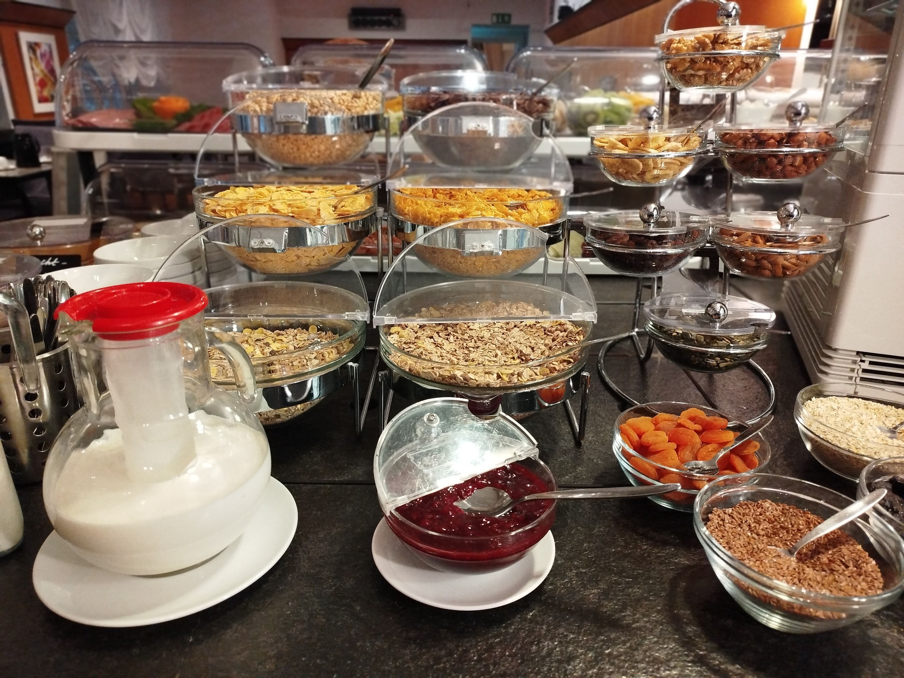 Buffet com diversos alimentos em recipientes transparentes, incluindo cereais, frutos secos e frutas desidratadas.