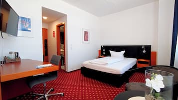 Habitación de hotel con cama, escritorio, silla, televisor y una pequeña mesa con un jarrón de flores.