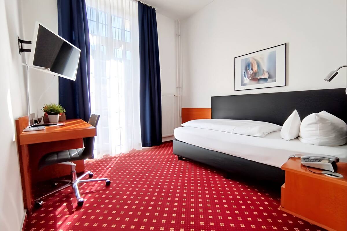 Habitación de hotel con cama, escritorio con computadora, una silla, lámpara de escritorio, planta y un cuadro en la pared.
