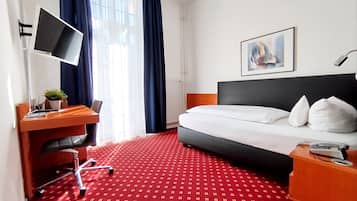 A hotel room with a bed, a desk with a computer, a chair, a desk lamp, a plant, and a painting on the wall.