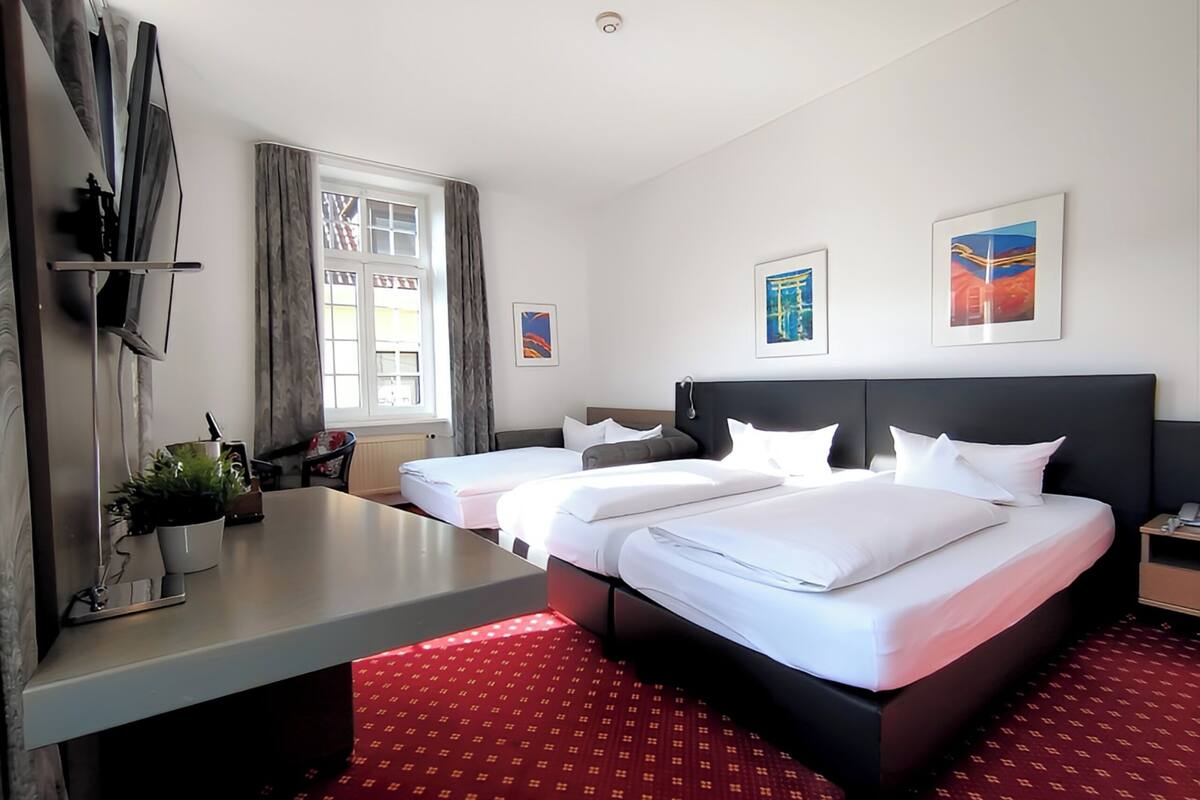 Habitación de hotel con dos camas, un escritorio, una ventana con cortinas y obras de arte en las paredes.