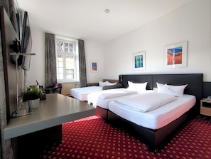 A hotel room with two beds, a desk, a window with curtains, and artwork on the walls. - Hotel Schwert (Rastatt)