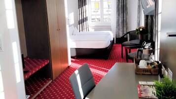 A compact hotel room with a bed, a desk, a chair, a small table, a TV, and a wardrobe.