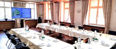 A conference room with long tables set for a meeting, equipped with chairs, tables, and a projector screen.