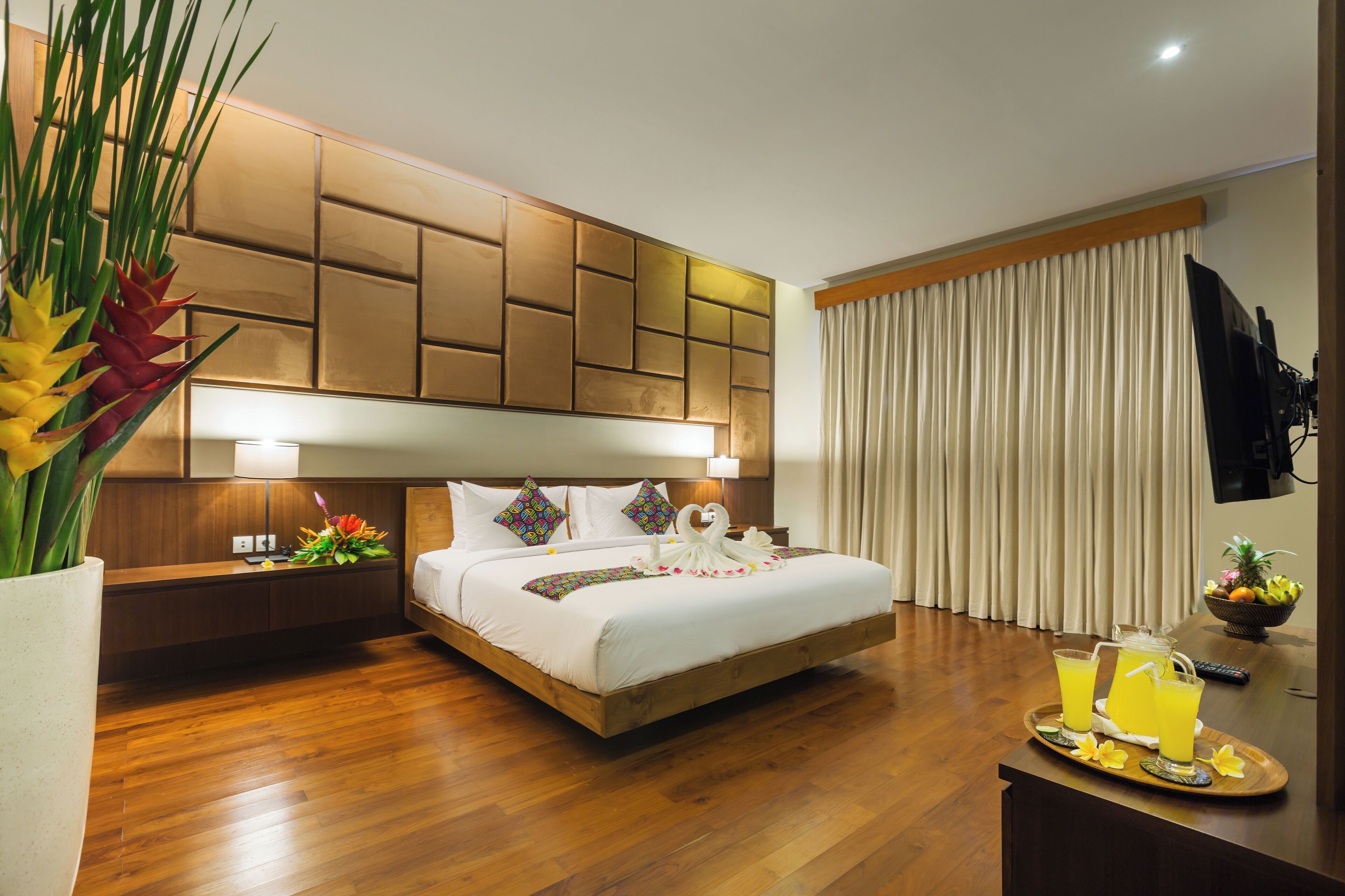 Habitación de hotel moderna con una cama de madera, una mesita de noche con una lámpara, una bandeja con bebidas y una planta grande en maceta.