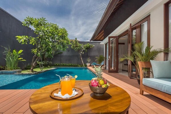 A poolside area with a wooden table, a fruit bowl, and a drink tray.