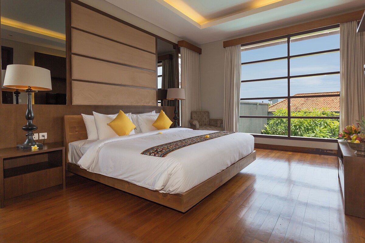 A spacious bedroom with a large bed, wooden flooring, a nightstand with a lamp, and a view of greenery through the window.