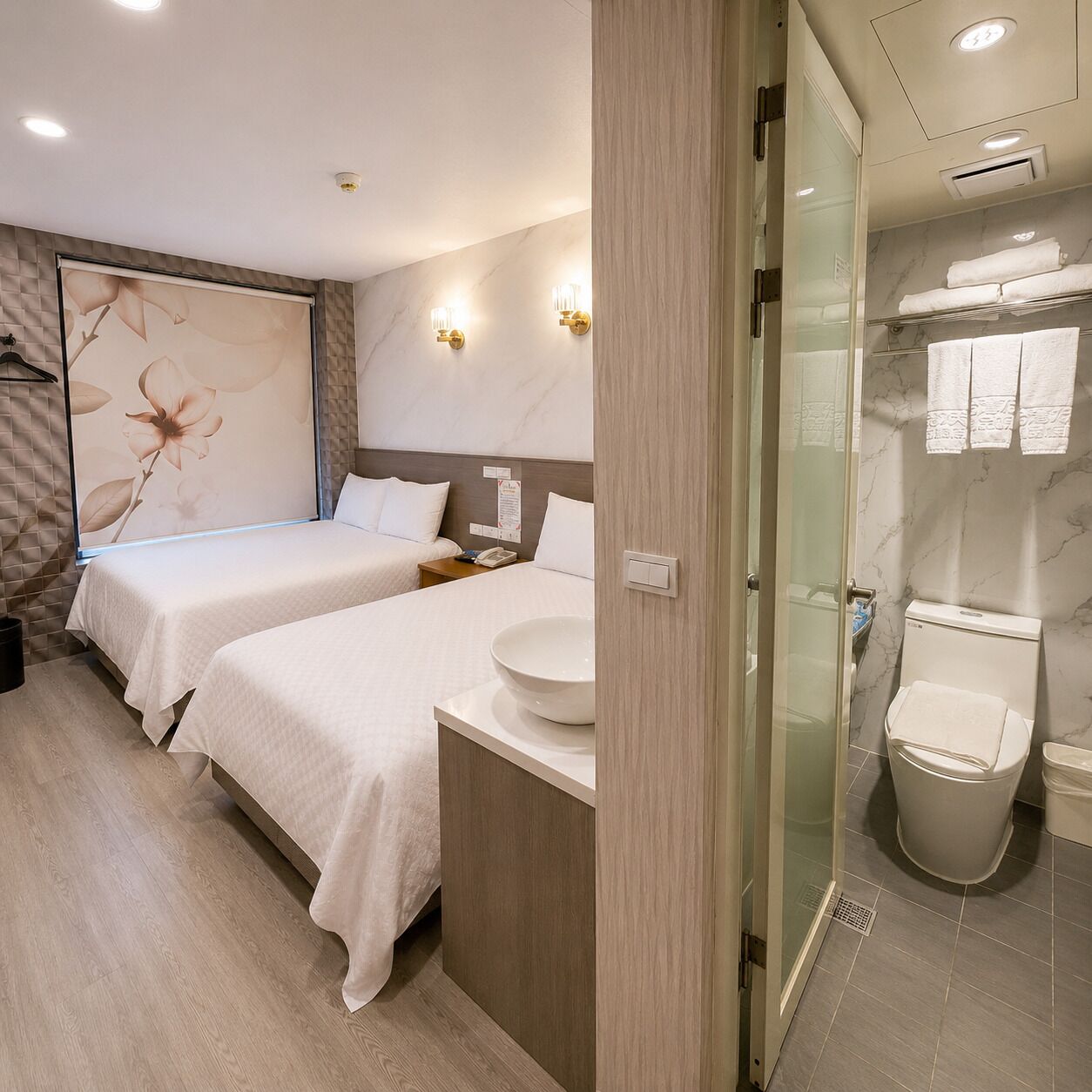 Quarto de hotel com duas camas, uma grande obra de arte na parede, banheiro com box de vidro e pia com piso de mármore.