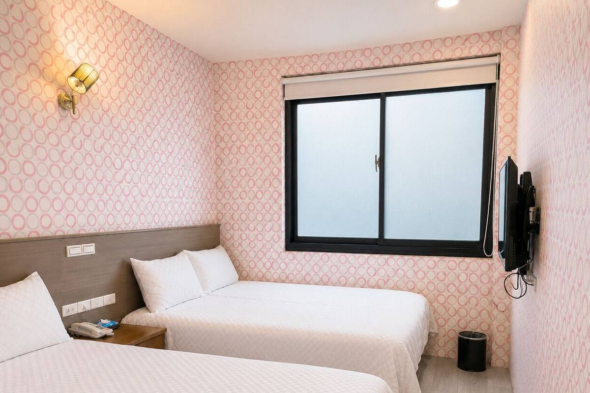 A hotel room with two beds, a TV, a window, and a wall-mounted light fixture.
