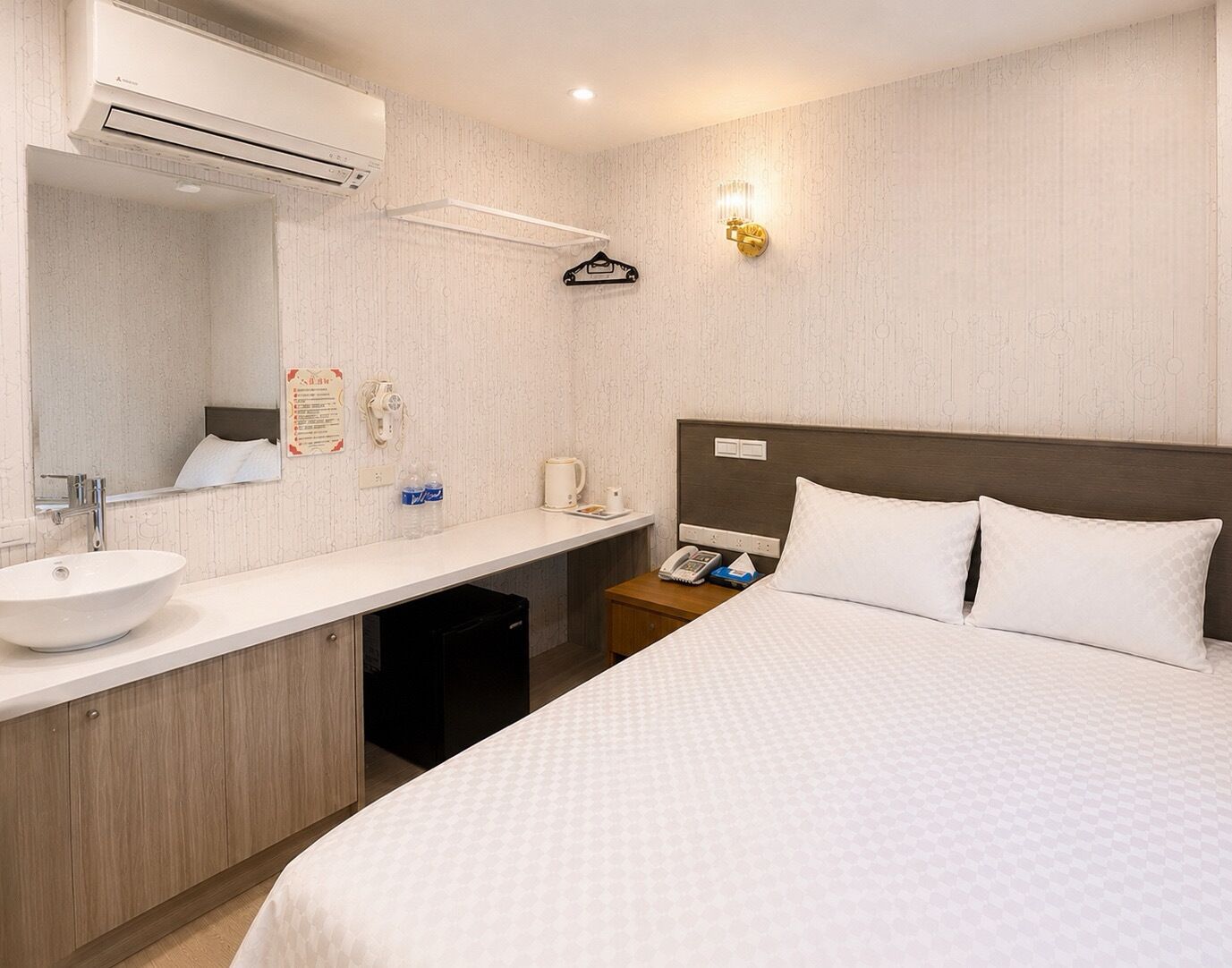 Habitación de hotel con cama, lavamanos, espejo, mesita de noche con teléfono y lámpara de pared.
