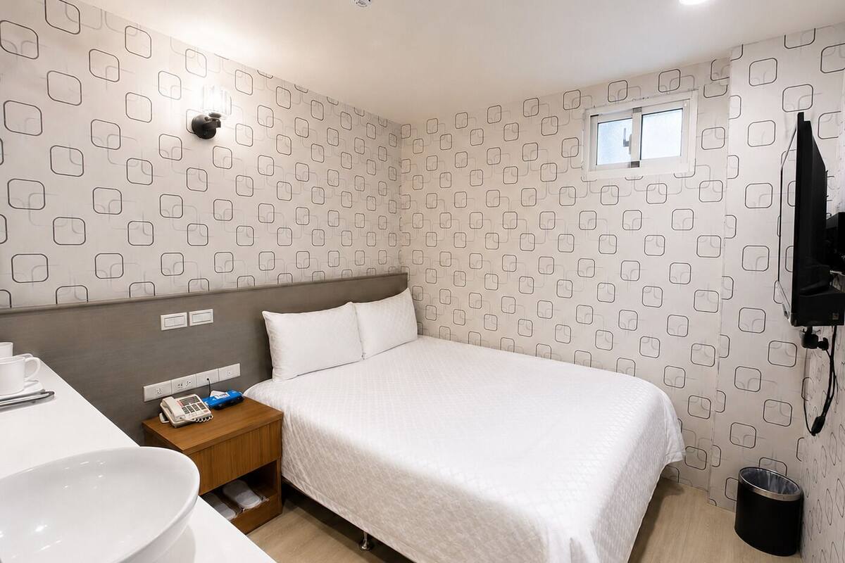 A hotel room with a bed, a bedside table with a phone and a cup, a sink, and a window.