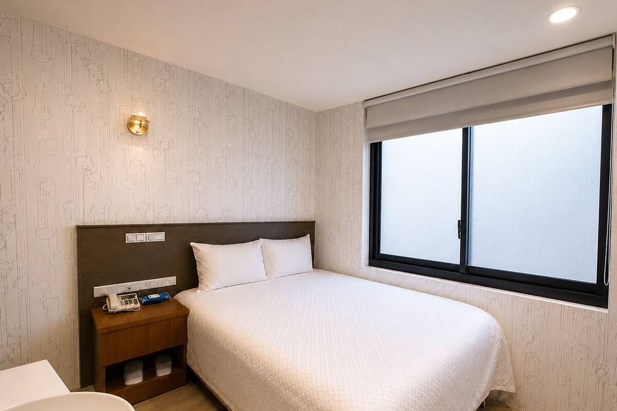 A hotel room with a large bed, a bedside table with a phone and a lamp, a window with blinds, and a bathroom with a sink and mirror.