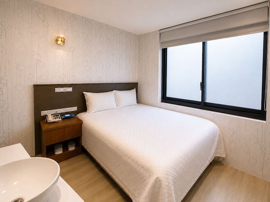 Quarto de hotel com uma cama grande, uma mesa de cabeceira com telefone e abajur, uma janela com persianas e um banheiro com pia e espelho.