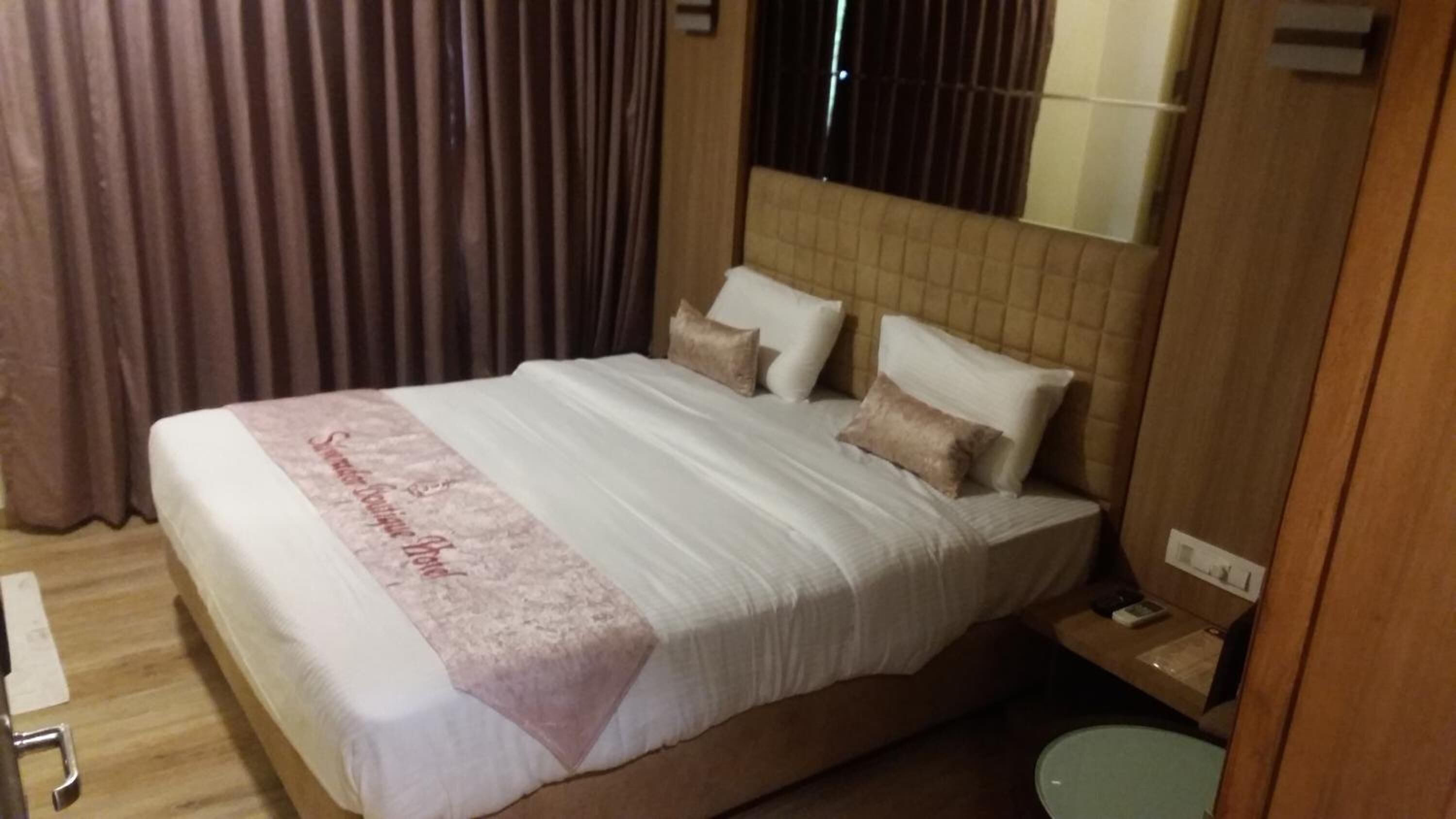 A hotel room with a large bed, a bedside table, a nightstand, and a window with curtains.
