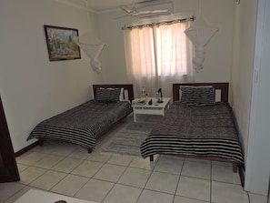 A room with two single beds, a nightstand with a lamp, a picture on the wall, and a window with curtains. - Hippo House Cottages (Victoria Falls)