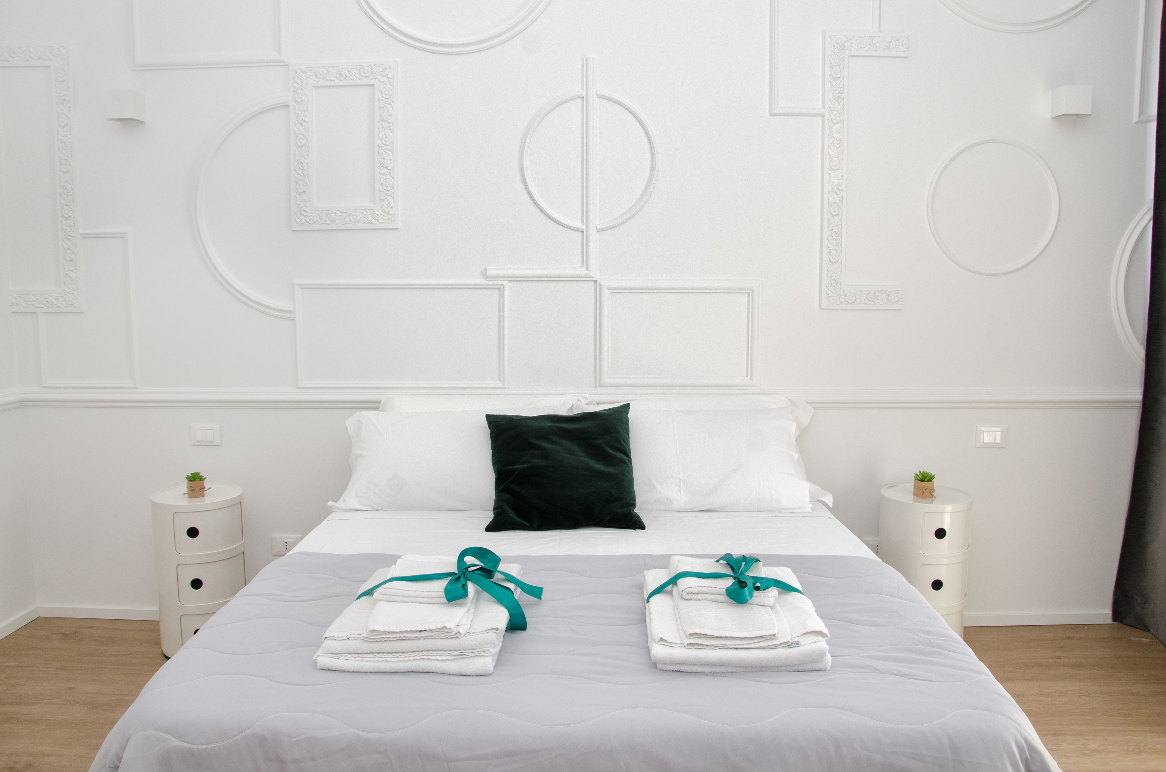 Una cama bien hecha con sábanas blancas y tiras verdes, dos mesitas de noche con pequeñas plantas y un almohadón verde oscuro sobre un cabecero blanco.