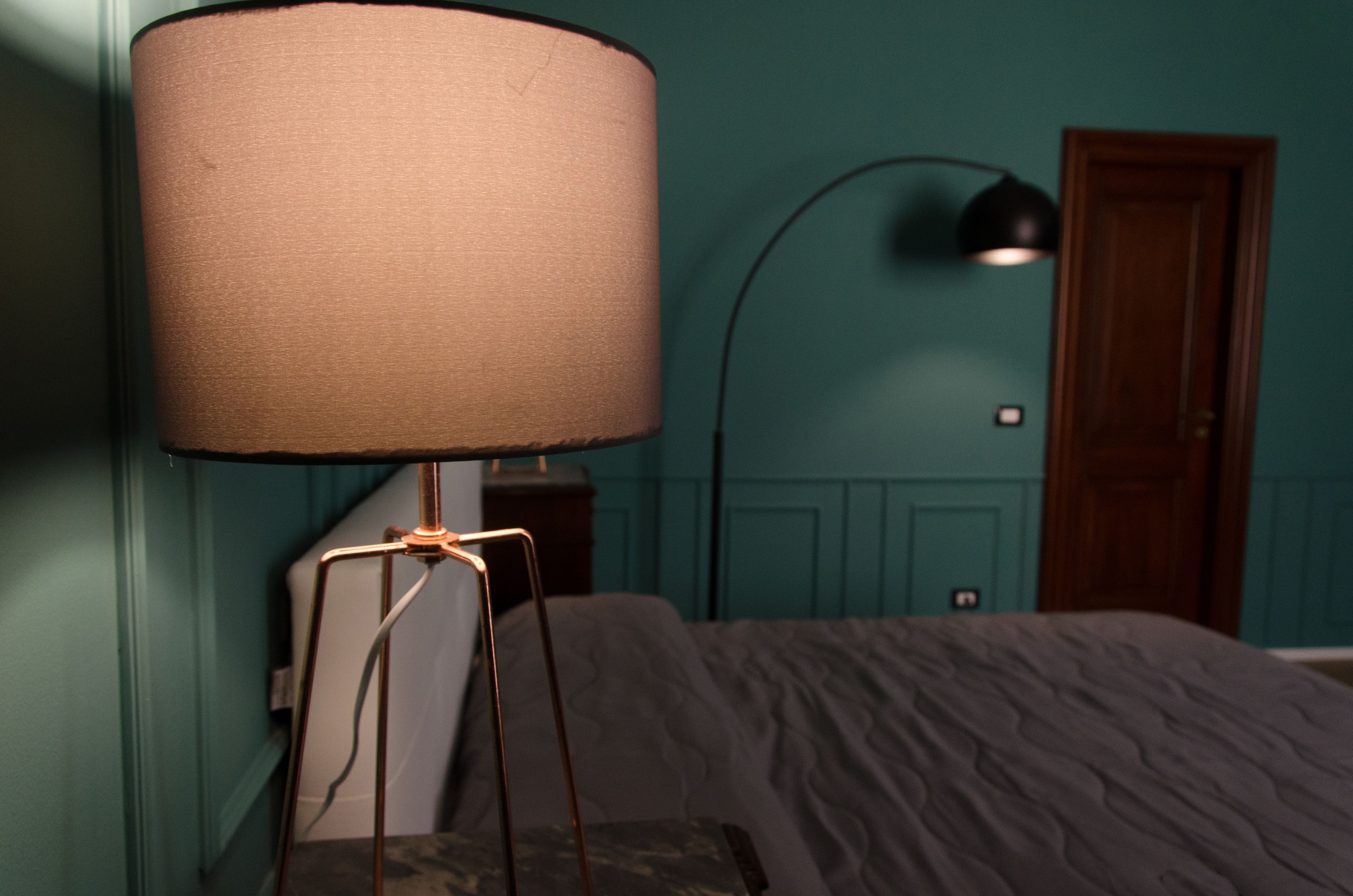 Una stanza dalle pareti verde acqua, con un letto con copripiumino a fantasia grigia, una lampada da terra con paralume beige e una lampada da tavolo nera.