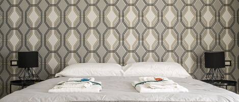 A double bed with white linens and towels, a geometric patterned wallpaper, and two bedside tables with lamps.