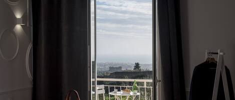 Una habitación con una cama, un sillón colgante, una mesa con una botella y vistas a un paisaje urbano a través de una puerta abierta.