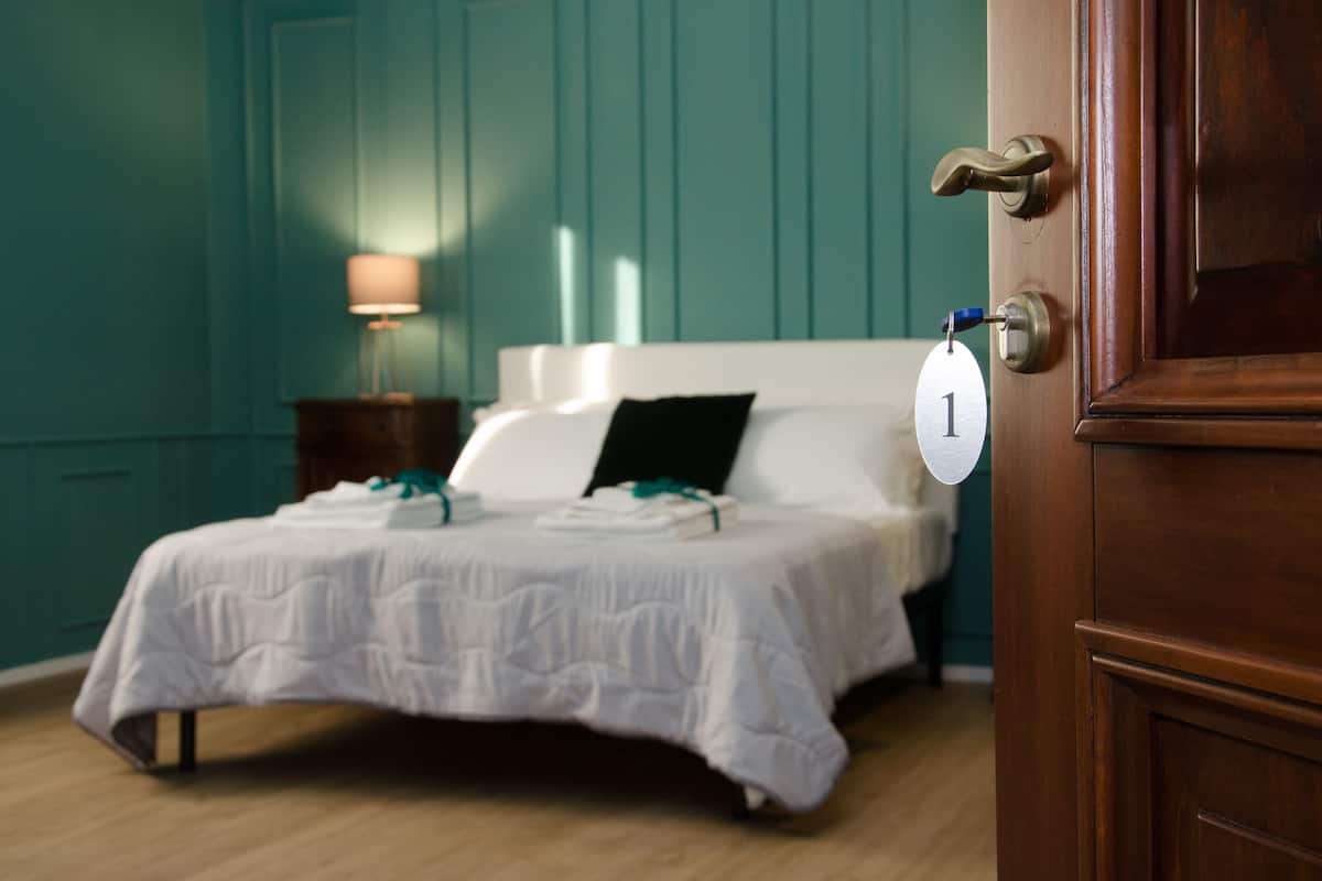 Una cama bien hecha con sábanas blancas, un almohadón negro y una mesita de noche con una lámpara. Se ve una puerta con el número 1.