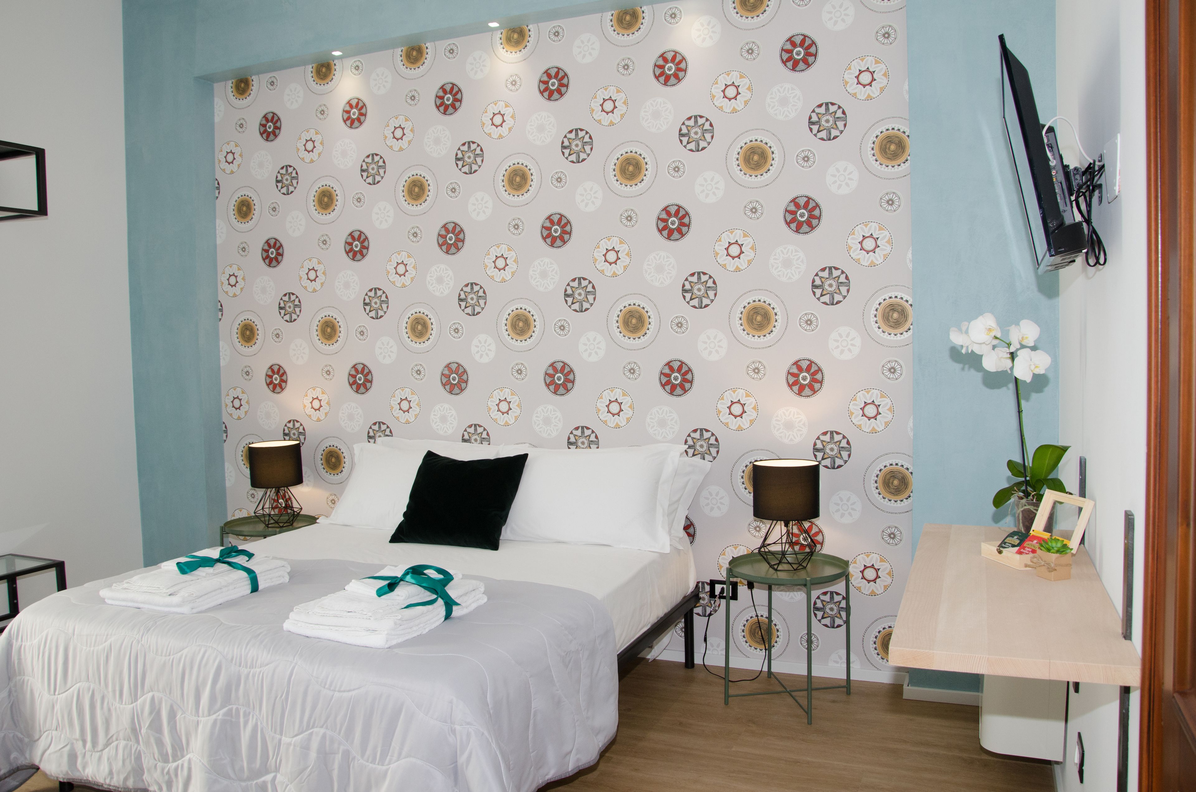 A neatly made bed with white linens and green towels, a wall-mounted TV, and a small table with a plant and a picture frame.