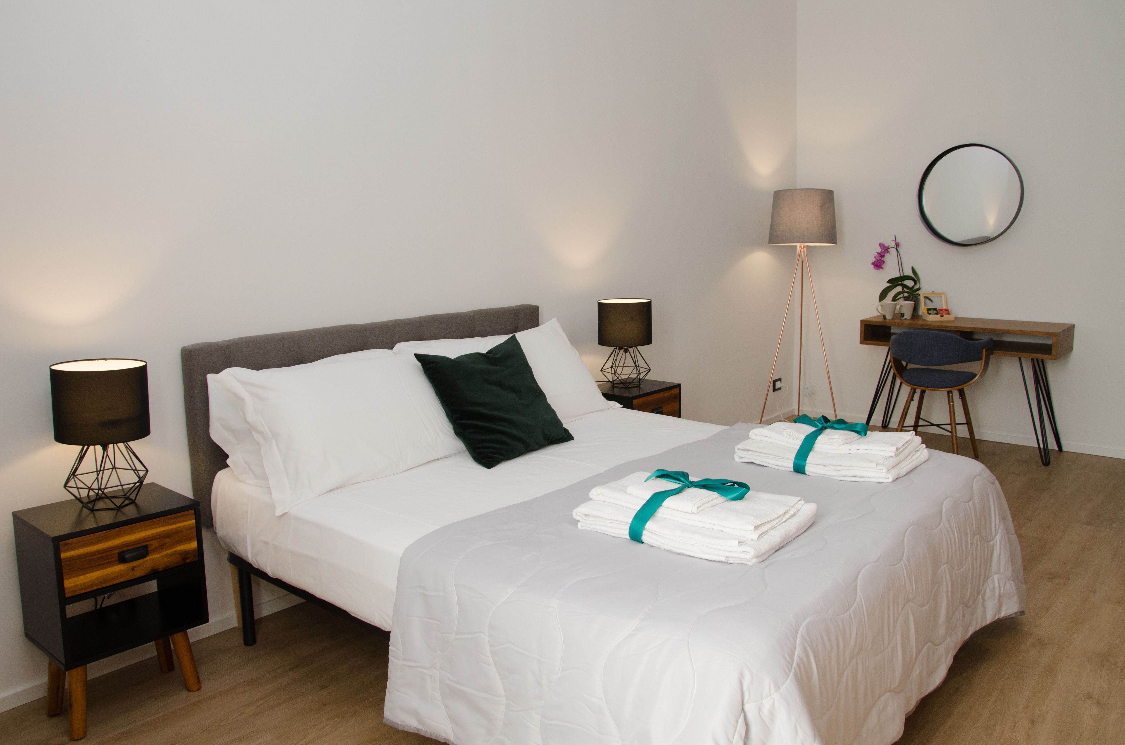 A neatly made bed with white linens and a teal ribbon, a bedside table with a lamp, a desk with a chair, and a mirror on the wall.