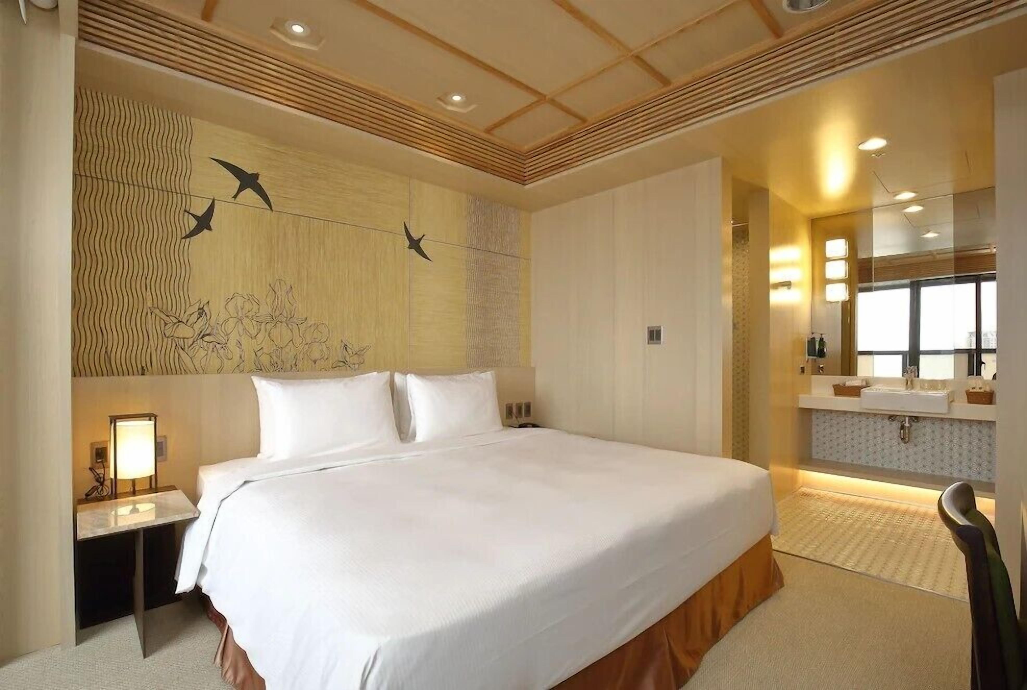 Une chambre d’hôtel avec un grand lit, une table de chevet avec une lampe, une salle de bain avec un lavabo et un miroir, et un mur décoratif orné d’oiseaux et de fleurs.