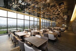 Un restaurante moderno con amplios ventanales que ofrecen vistas a la ciudad, mesas y sillas de madera, y un diseño de techo original con elementos colgantes.
