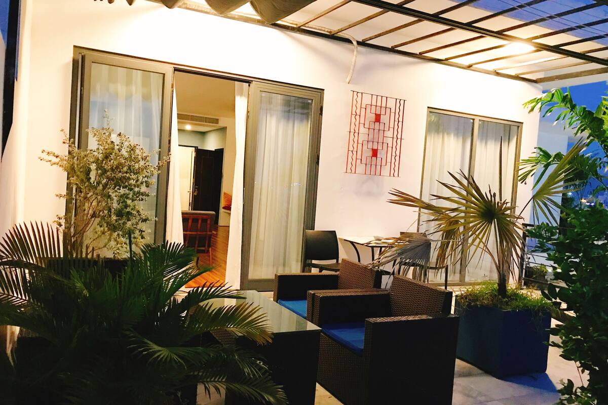 Un patio interior con mobiliario de mimbre, plantas en macetas y una mesa de comedor.