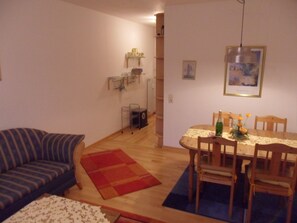 A living room with a sofa, a dining table with chairs, a red rug, and a wooden floor. - Strandleben 1 (Groemitz)