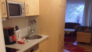 Una cucina compatta con microonde, bollitore e lavello. Il pavimento è in legno e c'è un tappeto a righe. Si intravedono un divano e una poltrona nella zona giorno adiacente.