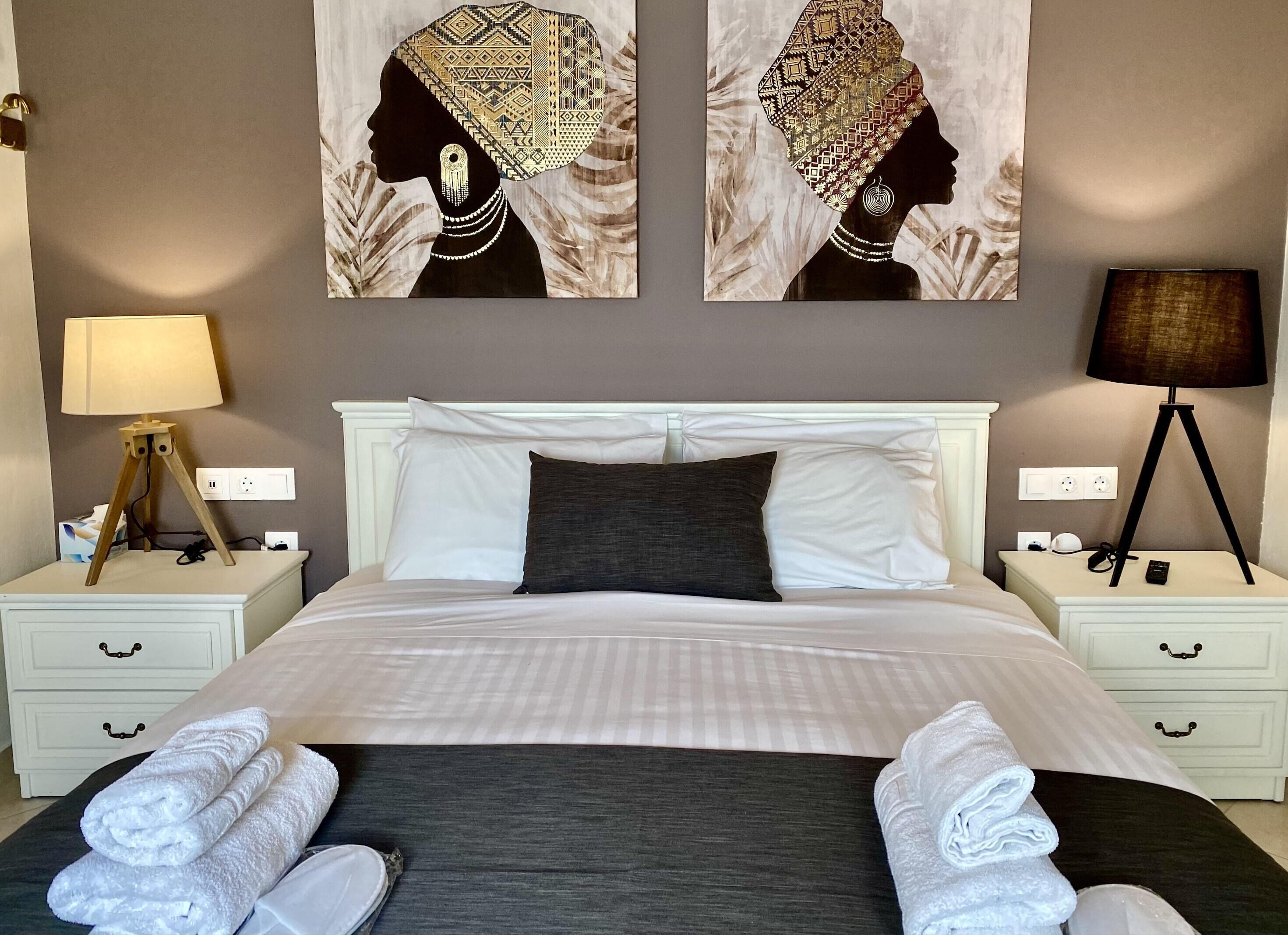 Cama bem arrumada com travesseiros brancos e escuros, uma luminária de cabeceira e uma luminária na parede. Duas mesas de cabeceira brancas com gavetas e um quadro grande acima da cama.