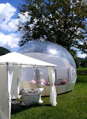 A transparent dome structure with a tent in front, a white table with a bottle labeled 'MOËT CHAMPAGNE', and a grassy field with trees in the background. - Bubble Tent Hotel (Weyregg am Attersee)