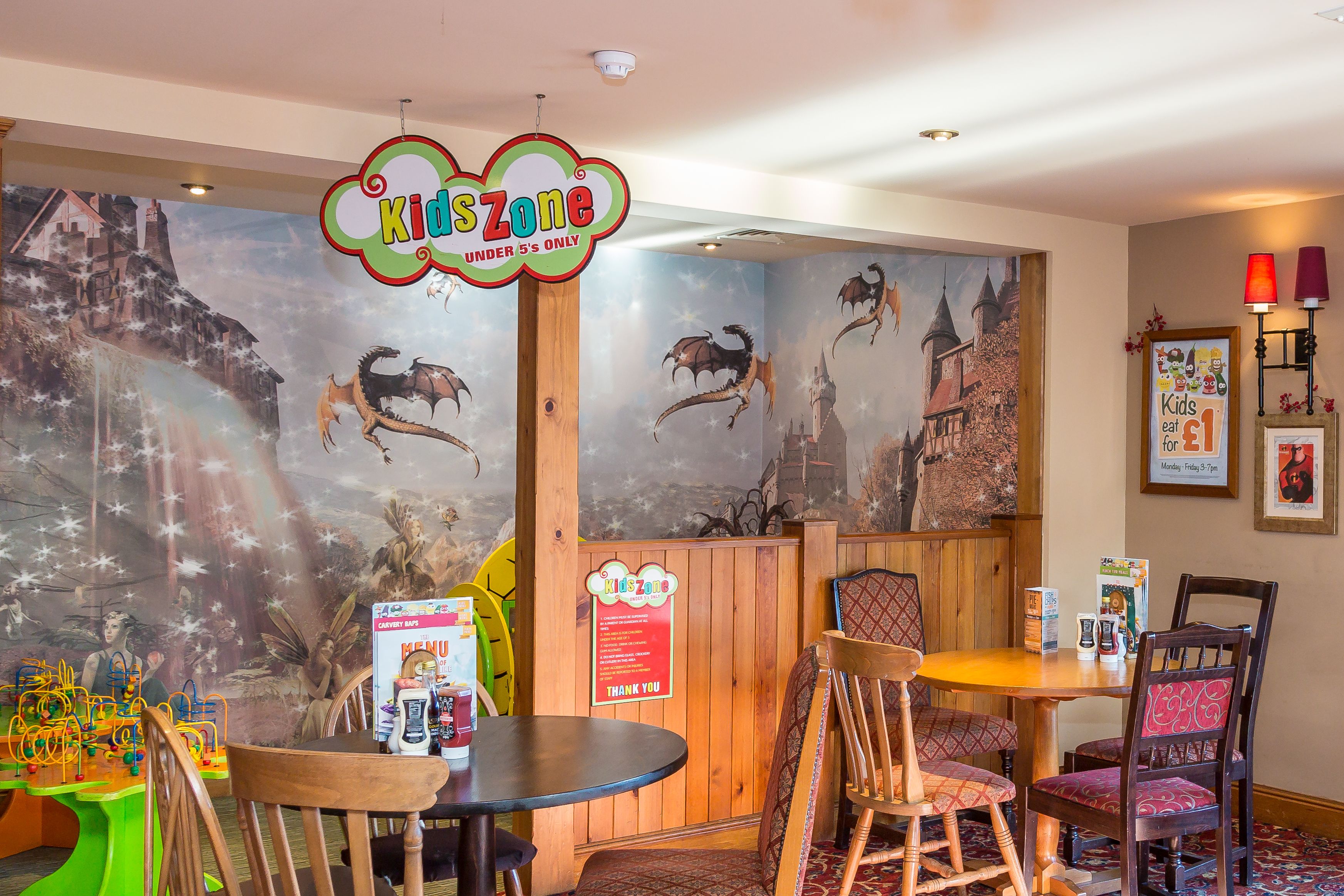 Restaurante infantil com placa 'KidsZone', móveis de madeira e murais com tema de dragões nas paredes.