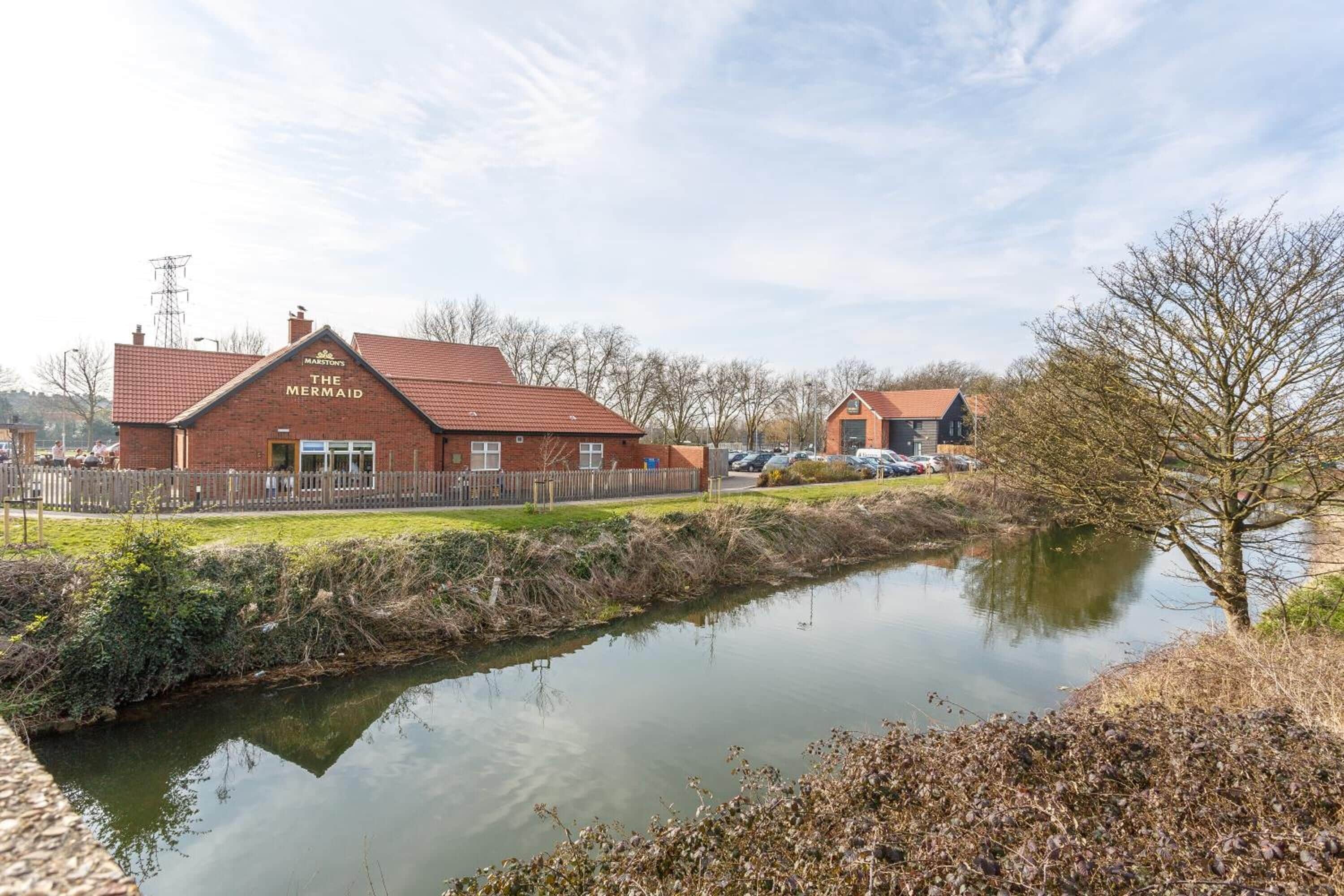 Un bâtiment en briques appelé « La Sirène », avec un toit rouge, situé à côté d’une étendue d’eau calme, entouré d’une végétation clairsemée et sous un ciel dégagé.