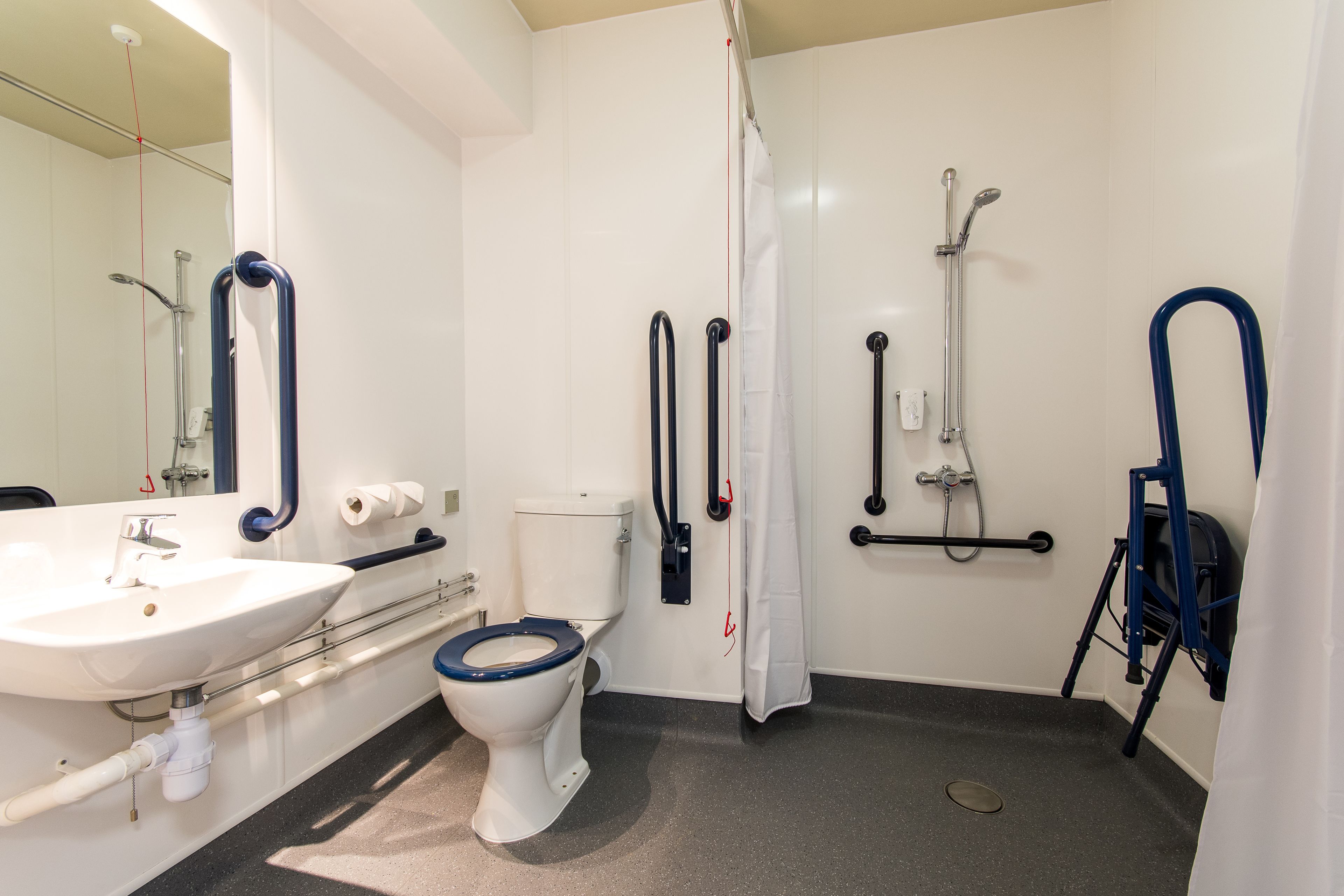 A bathroom with a white sink, a toilet with a blue seat, a shower with grab bars, and a wheelchair-accessible shower chair.