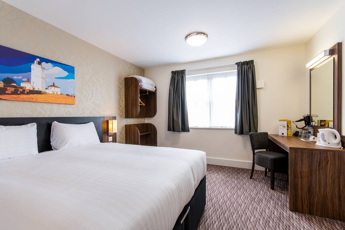 A hotel room with a large bed, a desk with a chair, a window with curtains, a painting of a lighthouse, and a mirror.