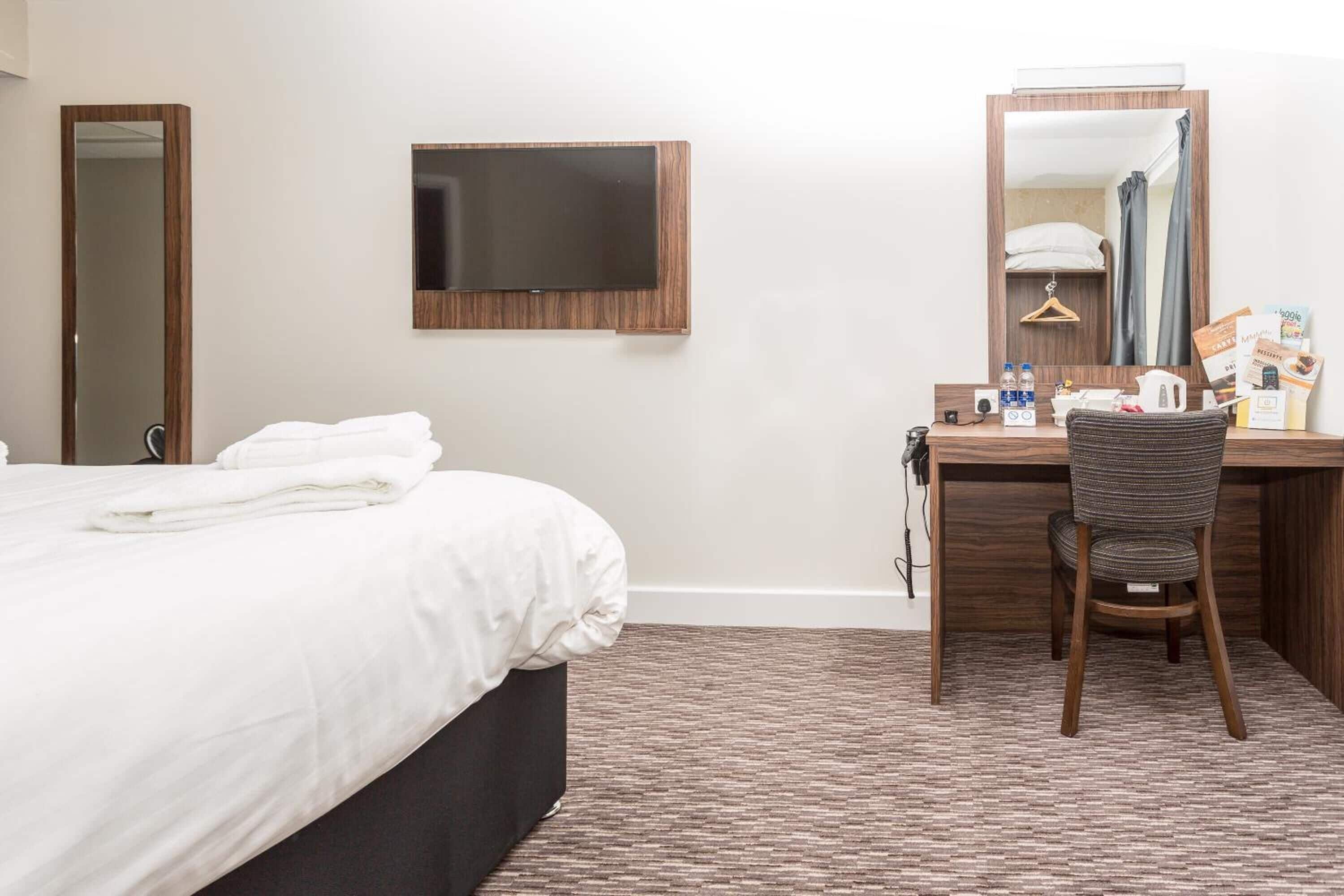 A hotel room with a bed, a desk with a chair, a flat-screen TV, and a mirror.