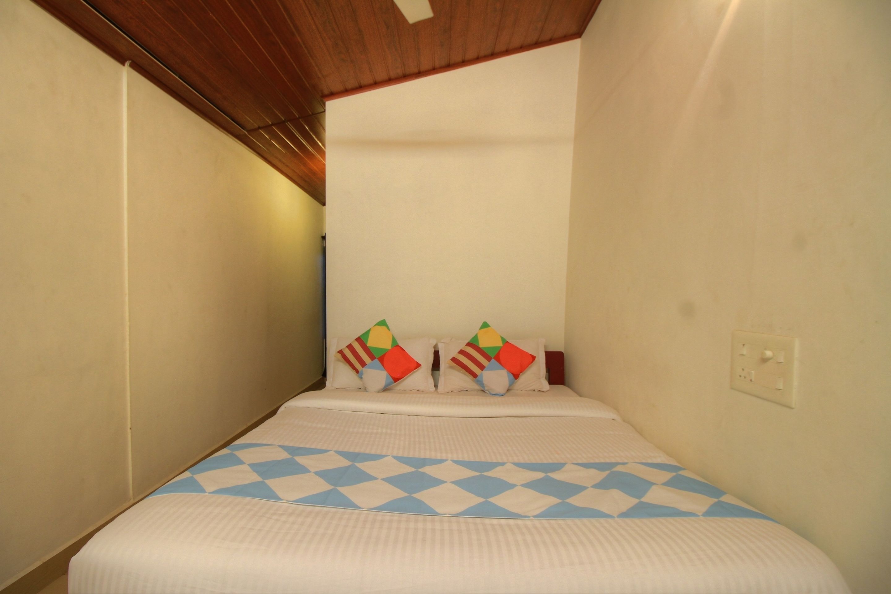 Een smalle hotelkamer met een enkel bed, een geruit sprei en twee kleurrijke kussens.