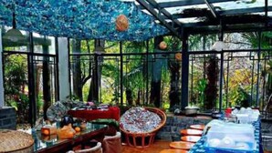 Uma área de refeições interna com uma mesa comprida coberta por um toalha azul, rodeada por cadeiras e bancos de madeira.