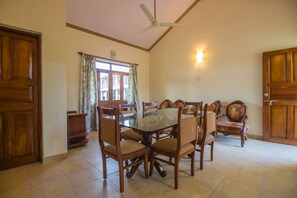 A dining area with a glass-top table surrounded by wooden chairs, a ceiling fan, and a window with curtains. - OYO 18705 Home Elegant 2BHK Parra (Arpora)