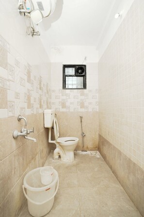 A small bathroom with a toilet, a shower, and a window. There are two buckets on the floor. - The Pearl Inn (Pune)