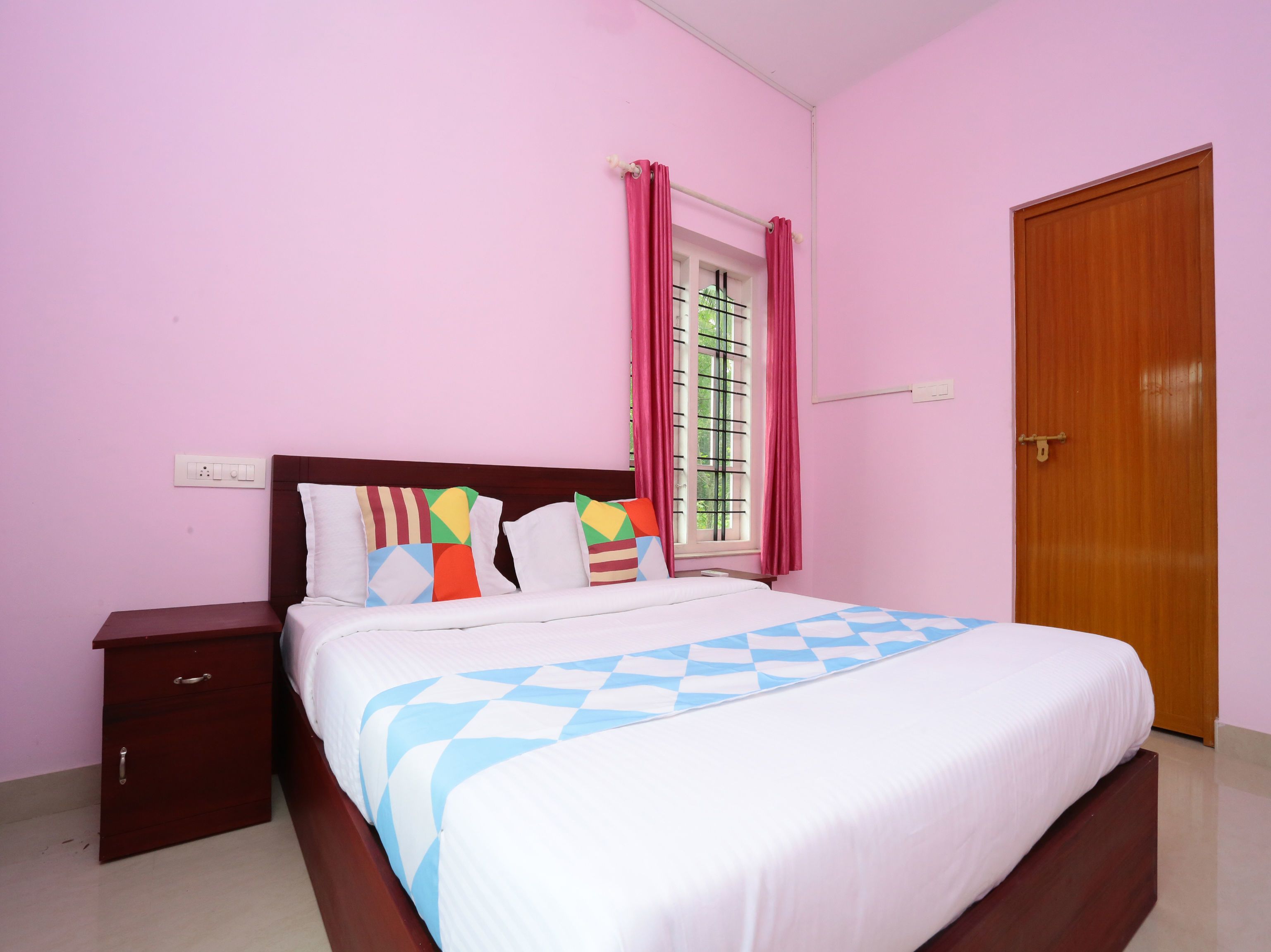 A bedroom with a bed, a nightstand, a wooden wardrobe, a window with curtains, and a wall socket.