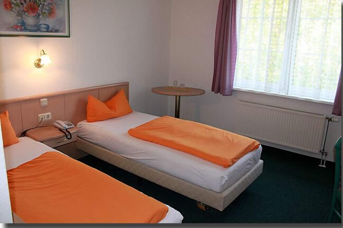 A hotel room with two beds, a bedside table with a lamp, a small round table, and a window with curtains.