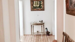 Un pasillo con suelo de madera, pared blanca, un cuadro enmarcado, una mesita con una figurita de pato y una silla.