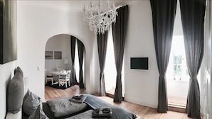 Un dormitorio moderno con una cama grande, cortinas oscuras y una araña. Se ve un comedor a través de un arco.