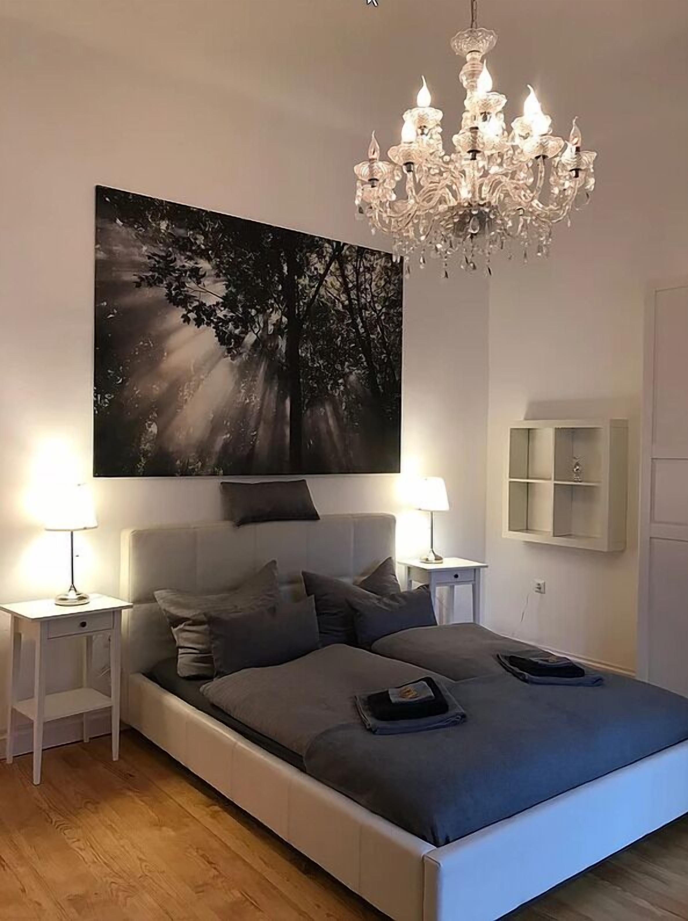 Ein Schlafzimmer mit einem großen Bett, zwei Nachttischen, einer Chandelüster, einem Wandcabinet und einem großen Wandbild mit Waldmotiv.