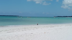 Een strand met helder turquoise water, boten in de verte en een persoon die over het zand loopt.