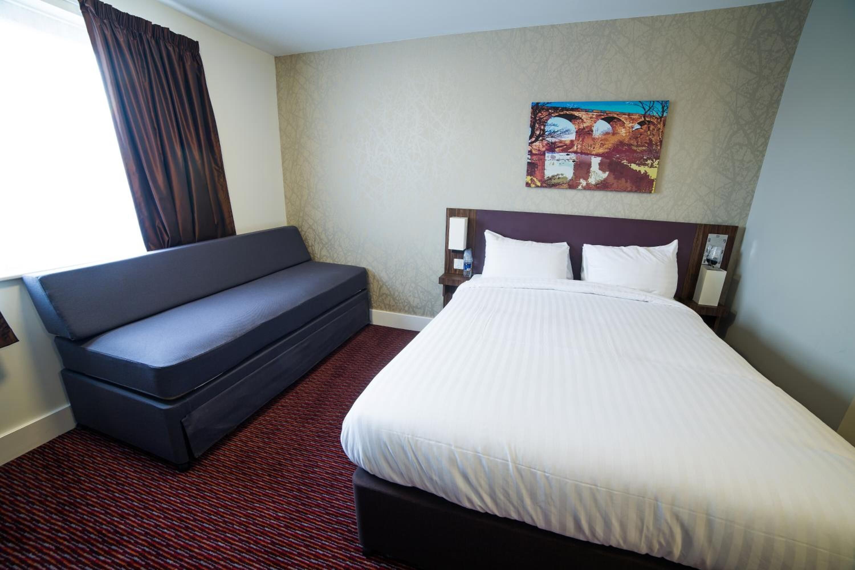 A hotel room with a bed, a sofa, a painting on the wall, and a window with curtains.