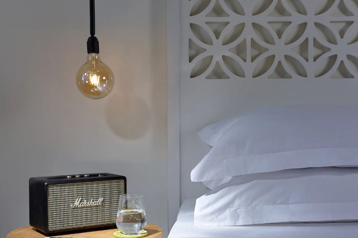 A bed with white linens, a Marshall speaker, a glass of water, and a hanging light bulb.